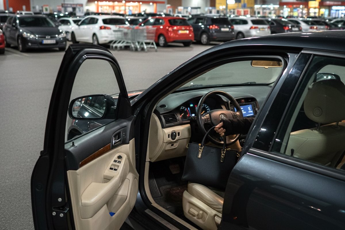 Interni di un'auto parcheggiata con portiera del conducente aperta, vista serale in un parcheggio di supermercato; truffa di una persona che ruba una borsa nera.
