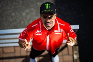Pilota di Formula 1 in giacca rossa e cappellino Monster Energy, sorridente e che fa il gesto della pace, seduto su una panchina.