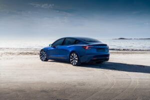 Auto elettrica Tesla Model 3 blu in spiaggia con vista sul mare al tramonto, veicolo innovativo e sostenibile, design moderno e tecnologia avanzata.