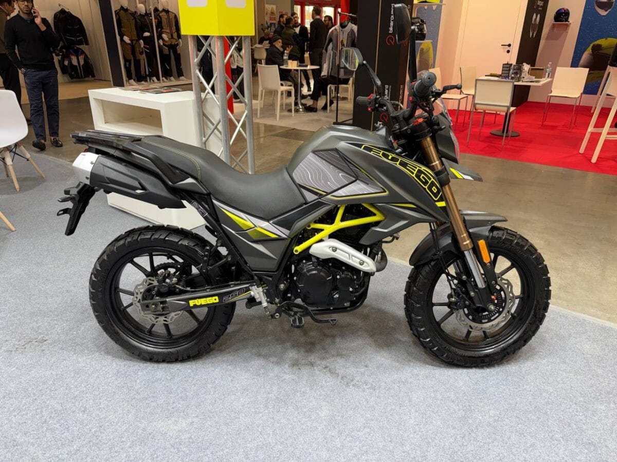 Moto enduro Fuego con telaio giallo e grafiche nere e grigie, esposizione in fiera, accessori e casco sullo sfondo.