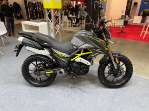 Moto enduro Fuego con telaio giallo e grafiche nere e grigie, esposizione in fiera, accessori e casco sullo sfondo.