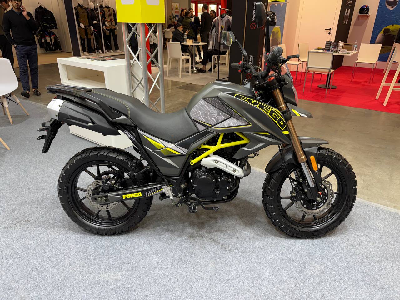 Moto enduro Fuego con telaio giallo e grafiche nere e grigie, esposizione in fiera, accessori e casco sullo sfondo.