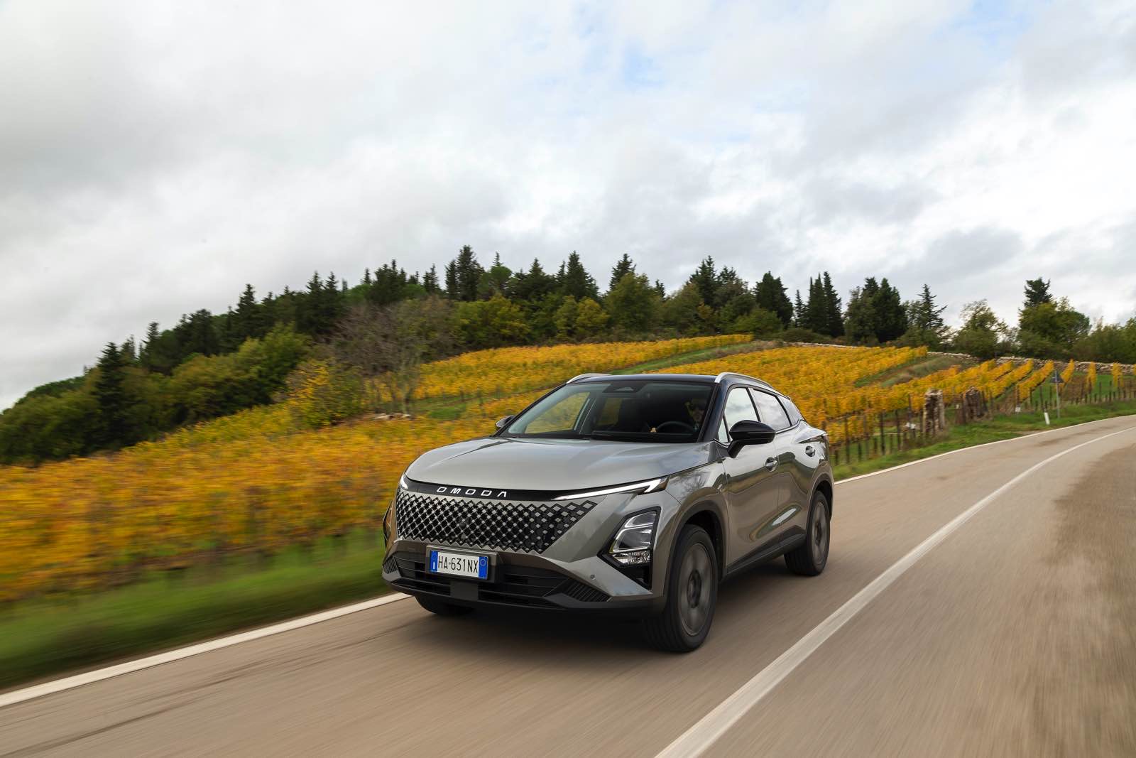 Nuova Omoda 5 SHS-S in movimento su strada panoramica con vigneti autunnali sullo sfondo, veicolo SUV moderno e dinamico.