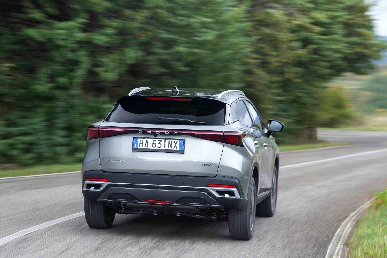 Auto SUV Omoda 5 SHS-S in movimento su strada panoramica circondata da natura, vista posteriore, design elegante e aerodinamico, tecnologia avanzata, veicolo ibrido, immagine dinamica e di lusso.