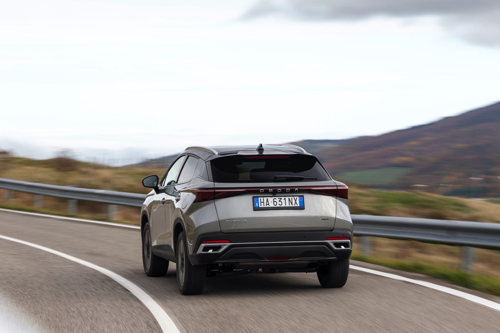 Auto SUV Omoda 5 SHS-S in movimento su strada di montagna con sfondo paesaggio naturale, immagine dinamica di veicolo elettrico, design futuristico, tecnologia avanzata, auto compatta di lusso, notizie automotive.