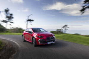 Auto Nuova Renault Clio 2026 rossa in azione su curva di strada panoramica, sfondo naturale con cielo nuvoloso, perfetta per immagini di veicoli sportivi e test drive.