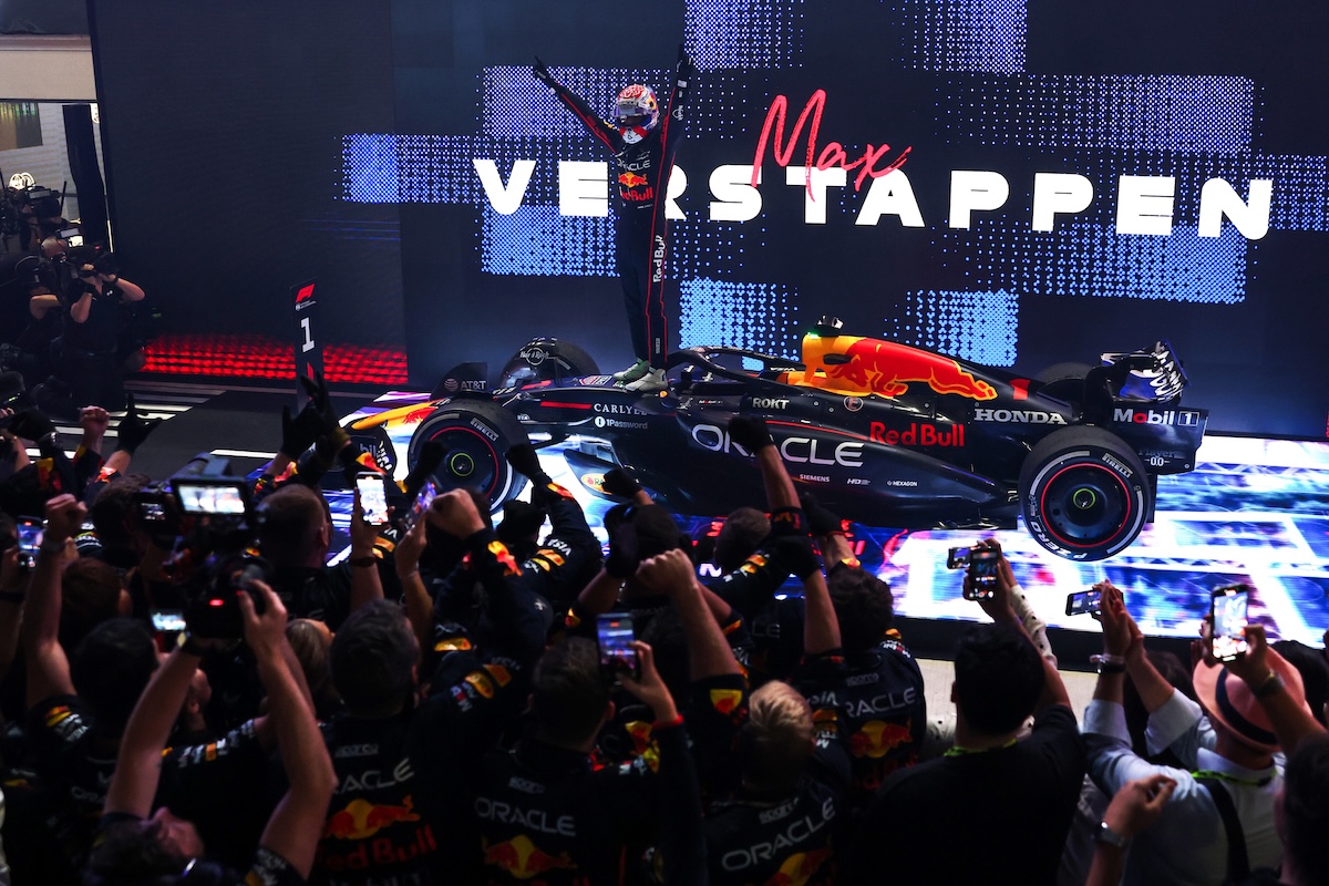 Descrizione: Max Verstappen celebra la vittoria in Formula 1 con il team Red Bull, sul podio durante un evento di gara, circondato da fan e media.