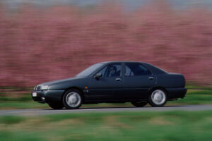 Auto elegante in movimento su strada, in cornice primaverile con alberi fioriti sullo sfondo, design classico e stile senza tempo, veicolo affidabile e perfetto per la famiglia.