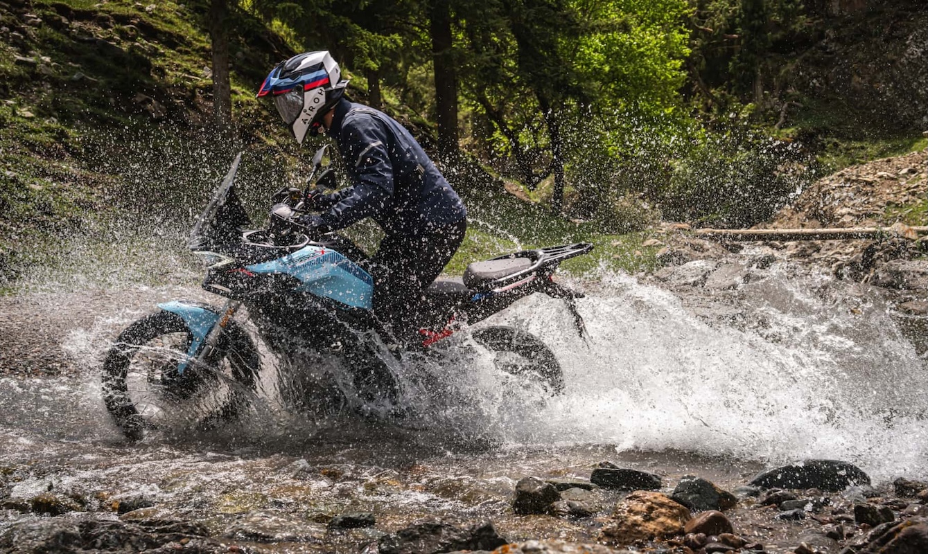 Motociclista con casco e abbigliamento sportivo su moto Morbidelli T352X attraversa un torrente in un'area naturale ricca di alberi.